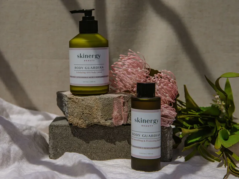 Olive-green lotion and serum bottles on stone blocks beside pink flowers and eucalyptus