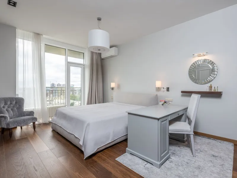 Bright modern bedroom with a made bed, grey armchair, and desk beside floor-to-ceiling windows