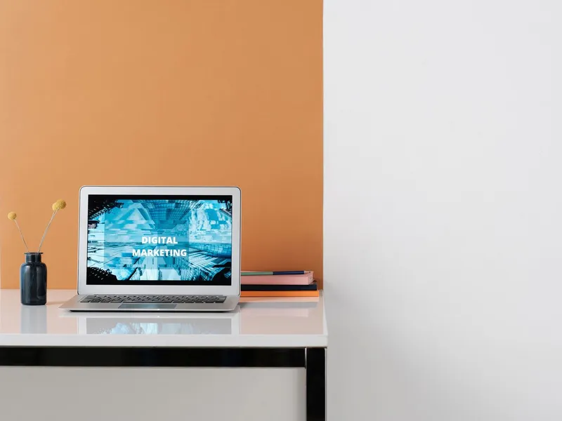 Silver laptop on a white desk in front of an orange wall, its screen reading “Digital Marketing”