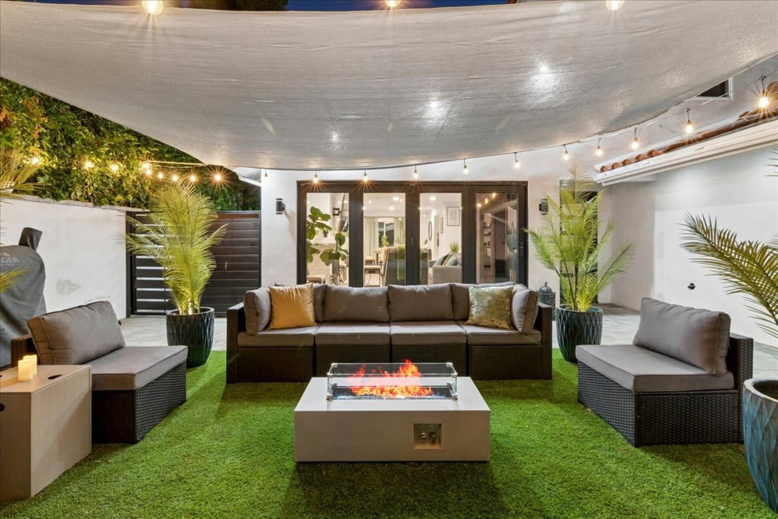 Backyard lounge with sectional seating around a lit fire table, under a shade sail and string lights