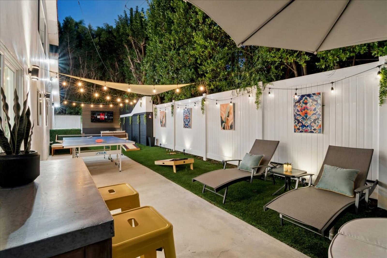Sun loungers along the backyard fence at dusk, with cornhole and ping pong beyond